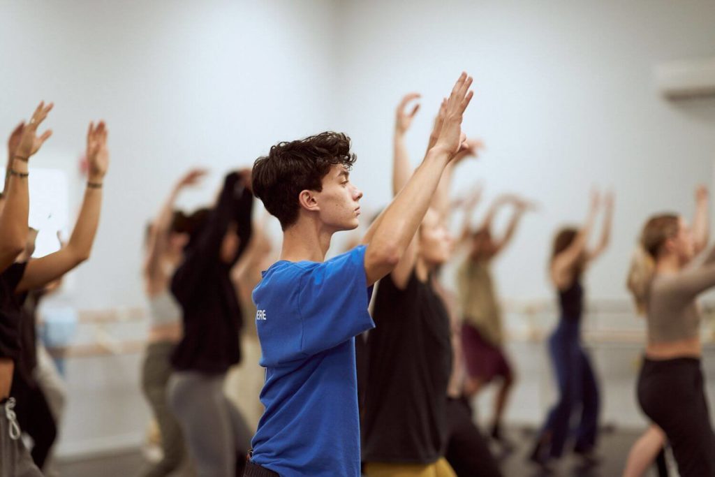 Dancer in blue shirt with hands above and in front of his head in a room full of other dancers at the CLI Conservatory