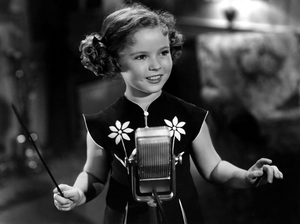 Shirley Temple holds baton and stands in front of a microphone wearing a dress with two flowers on it, smiling in black and white 