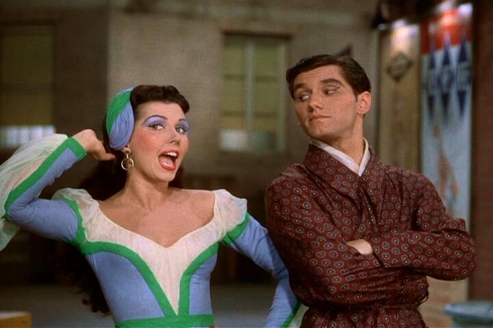 Ann Miller smiles wide wearing bright purple eyeshadow and dress with green and white details and a hat, next to man in red robe crossing his arms and looking at her