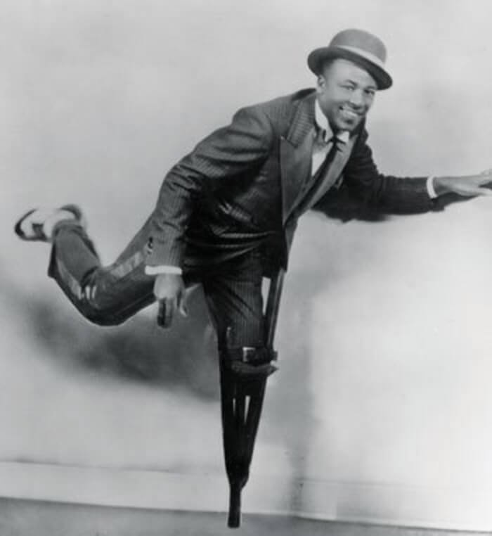 Clayton “Peg Leg” Bates wears suit bow tie and hat and balances on his peg leg, smiling and leaning over black and white