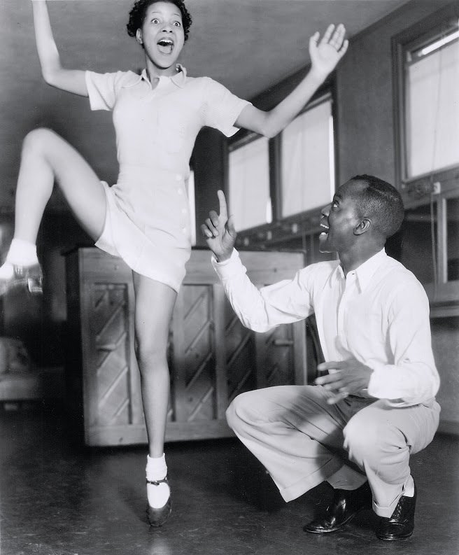 Jeni Le Gon in double deal. Black and white picture with Jeni Le Gon in tap shoes standing on one foot with a knee in the air and surprised expression. A man kneeling on the ground is next to her on her left.