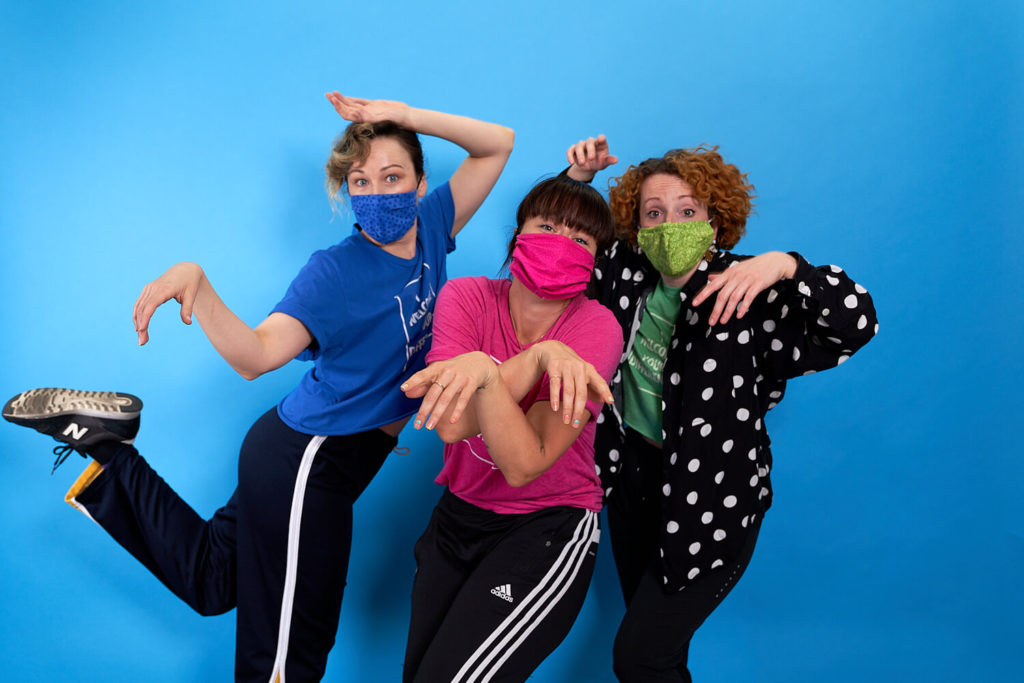 The Seaweed Sisters (Dana Wilson, Megan Lawson, Jillian Meyers) pose with green, pink, and blue masks and their hands posed by their heads