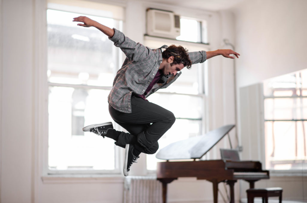 Ricky Ubeda jumping in the air with arms extended in front of grand piano in white studio.