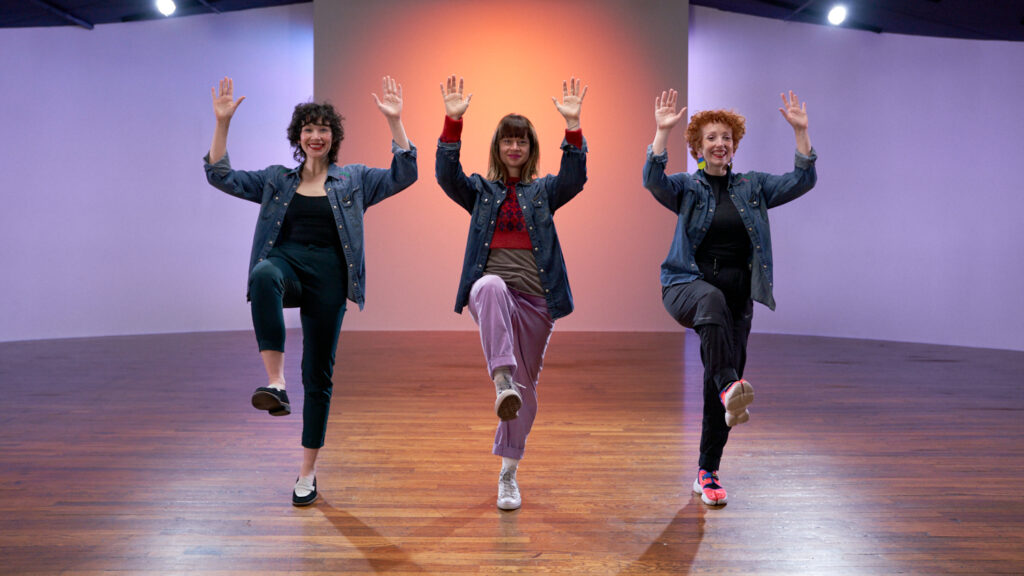 The Seaweed Sisters (Dana Wilson, Megan Lawson, Jillian Meyers) pose with their right leg lifted and their hands in the air.