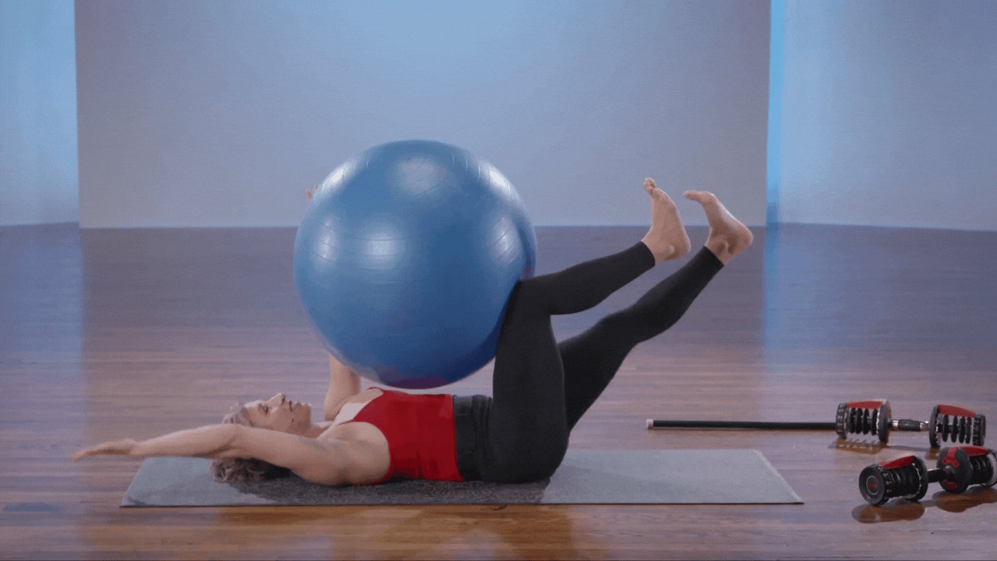Katie Schaar lying on her back with an exercise ball between her knees and arms demonstrating dead bugs extending one arm and one leg at a time.